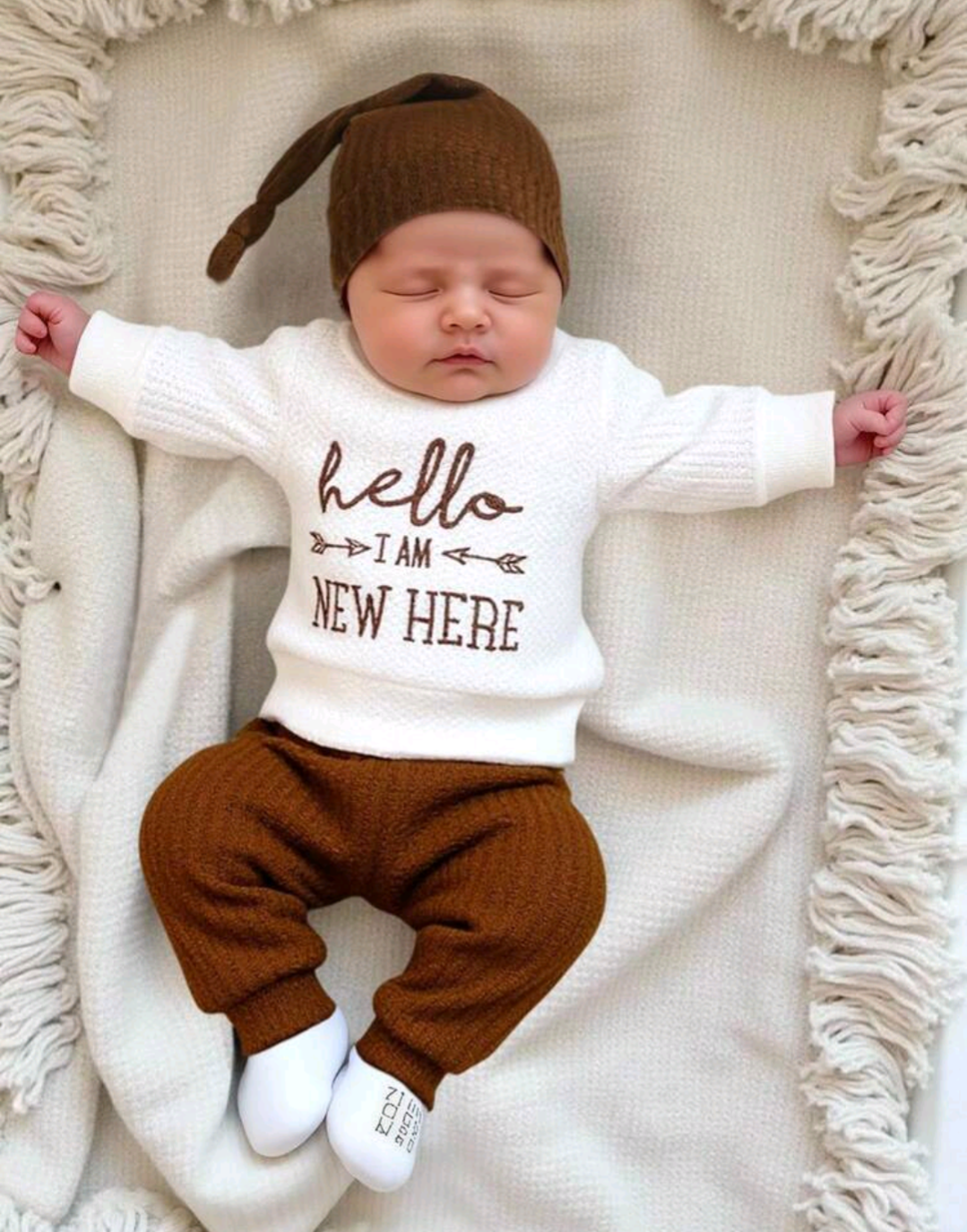 Newborn baby wearing white Hello I Am New Here knit sweater with brown ribbed trousers and knotted beanie hat
